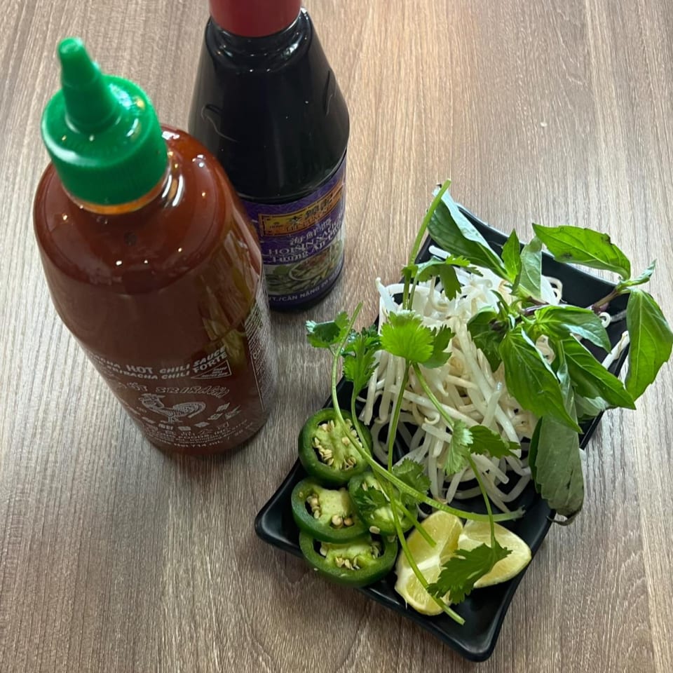 Sriracha and hoisin sauce bottles with a side plate of pho garnishes including bean sprouts, basil, cilantro, jalapeños, and lime.